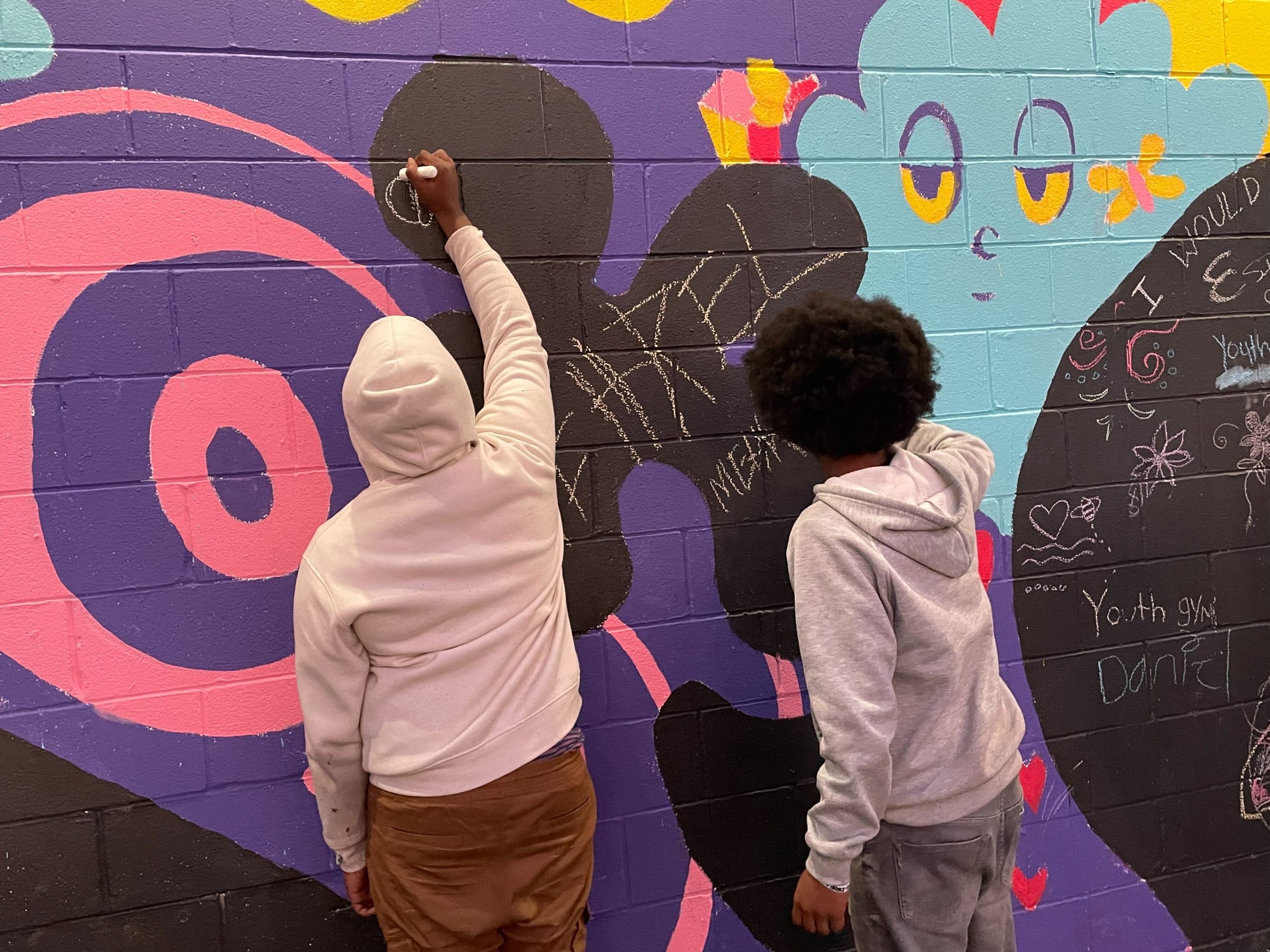 Two teenage boys draw with chalk on a blue, purple, black, and pink mural.